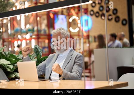 Glücklicher alter Senior Business man Investor, der sich ein Notebook ansieht, das den Gewinn feiert. Stockfoto