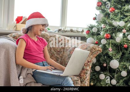 Ein glückliches kleines Mädchen öffnet zu Hause unter dem Weihnachtsbaum ein Geschenk und kommuniziert mit ihren Großeltern über einen Laptop via Videolink. Urlaubskonzept Stockfoto