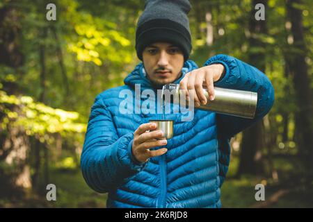 Wanderer auf einem langen Weg ruhen sich aus und geben heißen Tee aus einer Thermoskanne in einen Becher, um sich aufzuwärmen und Vitamine und Flüssigkeiten aufzufüllen. Hochgebirgswandern. Stockfoto