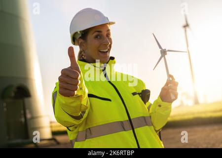 Eine Ingenieurin mit einem Windturbinenmodell und einem Windpark mit Daumen nach oben. Bild des neuen Energiekonzepts Stockfoto