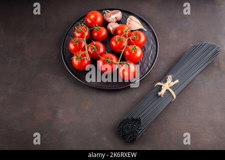 Trockene schwarze Pasta-Spaghetti, Tomaten und Knoblauch auf dunklem Hintergrund. Schwarze Spaghetti mit Seil gebunden und frisches Gemüse auf einem schwarzen Teller. Draufsicht. Cop Stockfoto