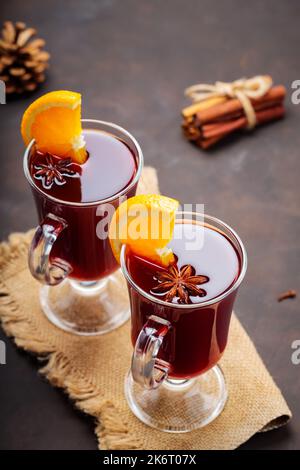 Glühwein mit Orangenscheiben und Gewürzen auf Sacktuch. Heißer Glühwein und Zutaten auf dunklem Hintergrund. Wärmendes Herbstgetränk-Konzept Stockfoto