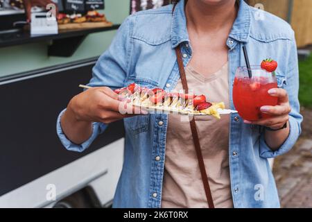 Die Frau genießt köstliche Pfannkuchen mit Erdbeeren und Erdbeer-Daiquiri auf einem Food Festival. Crepes mit frischen Erdbeeren und Schlagsahne, Street Stockfoto