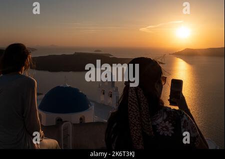 Touristen, die Fotos machen und den Sonnenuntergang mit den drei Glocken von Fira beobachten, offiziell bekannt als die katholische Kirche der Dormition, mit ihrem trad Stockfoto