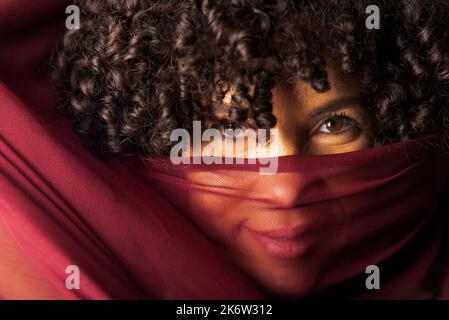 Porträt einer schönen Frau mit einem dünnen Tuch von dunkelroter Farbe auf ihrem Gesicht vor schwarzem Hintergrund. Reife Frau. Selbstwertgefühl-Konzept. Stockfoto
