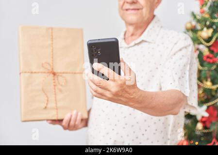 Ein älterer Mann zeigt in einem Videoanruf das Weihnachtsgeschenk, das er gerade erhalten hat. Konzepte: Die Freude am Teilen im Urlaub, der Einsatz von Technologie und Soziales Stockfoto