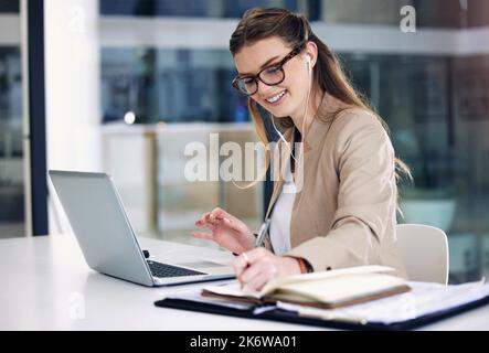 Shes eine fleißige Biene wie immer. Eine junge Geschäftsfrau, die Ohrhörer trägt, während sie Notizen schreibt und in einem Büro an einem Laptop arbeitet. Stockfoto