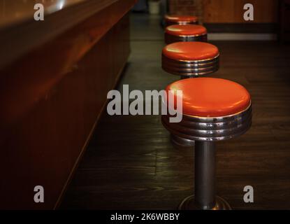 Vintage Hocker im Restaurant. Reihen Sie Vintage-Hocker vor einer Holztheke in einer Bar im Vintage-Stil. Eine Reihe von vier glänzenden roten Vinylhockern. Nobo Stockfoto