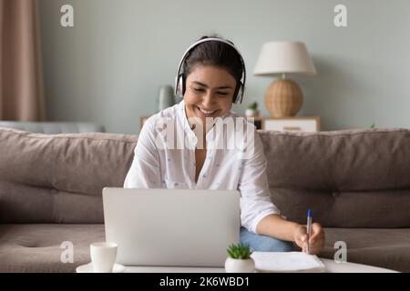 Fröhliche indische Frau in Kopfhörern mit Laptop zu Hause lernen Stockfoto