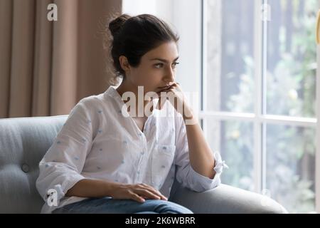 Traurige Inderin sitzt auf dem Sessel sieht besorgt aus, fühlt sich verwirrt Stockfoto