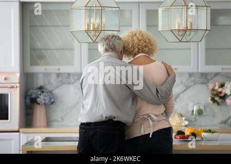 Rückansicht liebevoller älterer Mann umarmte Frau beim Zubereiten des Frühstücks in der Küche Stockfoto
