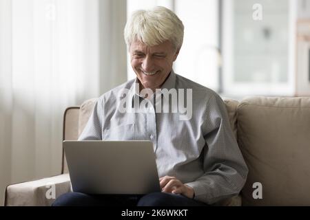 Glücklicher älterer Mann sitzt auf dem Sofa und benutzt einen modernen Laptop Stockfoto