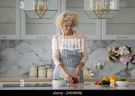 Ältere Frau in Schürze schneidet roten Paprika bereiten vegetarischen Salat Stockfoto