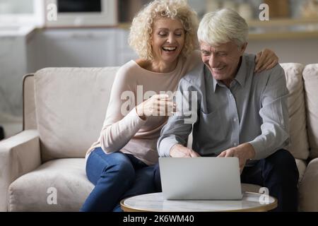 Ein älteres Paar sitzt lachend auf dem Sofa und starrt auf den Laptop-Bildschirm Stockfoto