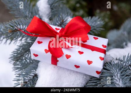 Eine weiße Schachtel in Herzen mit einer roten Schleife auf einem Zweig eines Weihnachtsbaums und fallendem Schnee. Das Konzept der Feier des Valentinstag oder Geschenke zum Valentinstag, Geburtstag oder anderen Feiertagen. Hochwertige Fotos Stockfoto