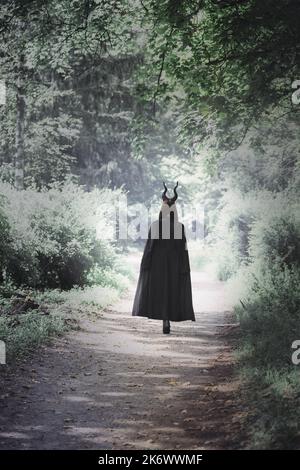 Eine Hexe mit langen Hörnern im dunklen Mantel geht durch einen dunklen, gespenstischen Wald. Halloween-Konzept Stockfoto