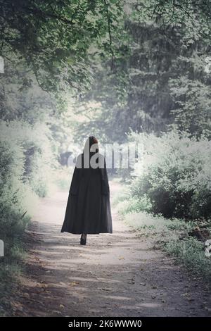 Eine Frau in einem schwarzen Cape geht durch einen dunklen, gruseligen Wald. Halloween-Konzept Stockfoto