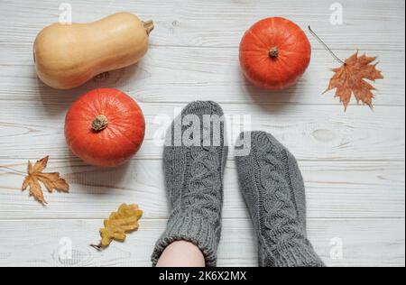 Beine eines Mädchens in gestrickten Socken auf einem Holzhintergrund neben Kürbissen und Herbstblättern. Gemütliches Herbstkonzept. Stockfoto