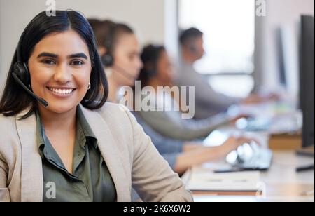 ID freut sich, Ihnen zu helfen. Porträt einer jungen Callcenter-Agentin, die in einem Büro arbeitet, mit ihren Kollegen im Hintergrund. Stockfoto