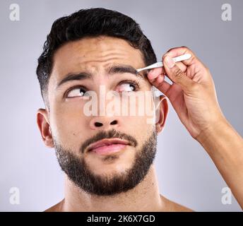 Ich habe es eher einfach mit überholen. Nahaufnahme eines jungen Mannes, der seine Augenbrauen vor grauem Hintergrund zupft. Stockfoto