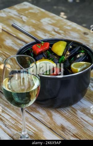 Ein Glas Weißwein vor einem Gericht mit gekochten Muscheln in Muscheln mit Zitronenscheiben auf einem Holztisch aus der Perspektive. Stockfoto