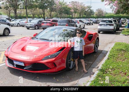 Atibaia - Brazil, October 7, 2022: Boy in sunglasses posing next to a parked red Ferrari F8 Tributo. Mid-engined rear drive sports car. Ferrari - Ital Stockfoto