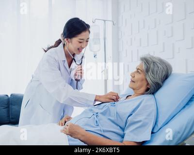 Eine schöne junge asiatische Ärztin in weißem Anzug lächelte, während sie Stethoskop benutzte, um zu untersuchen und dem Herzschlag einer älteren alten Patientin in blu zu lauschen Stockfoto
