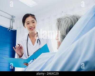 Junge asiatische Ärztinnen besuchen eine ältere Patientin im Bett und erklären die Symptome und Untersuchungsnotizen sowie medizinische Gesundheitsdokumente auf Clipbo Stockfoto