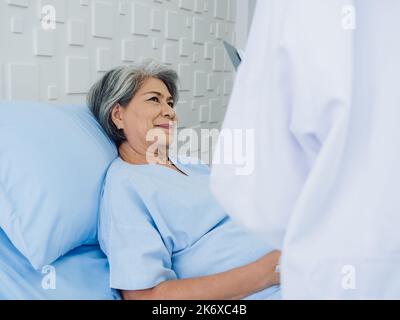 Lächeln Asiatische ältere Frau graue Haare Patientin auf dem Bett liegend, erholt sich, während junge Ärztinnen besuchen, erklären die Symptome und Untersuchungsnotizen ein Stockfoto