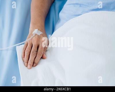 Nahaufnahme der Kochsalzlösung bei älteren Patienten Hand in blauem Kleid, das mit weißer Decke im Krankenhauszimmer mit Kopierraum auf dem Bett liegt. Ha des älteren Patienten Stockfoto