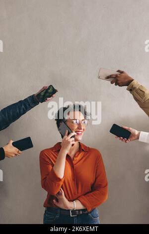 Glückliche Geschäftsfrau, die am Telefon spricht, während sie von Händen umgeben ist, die Smartphones halten. Fröhliche Geschäftsfrau, die lächelt, während sie Anrufe entgegennimmt Stockfoto