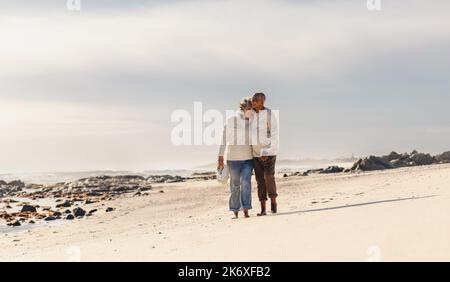 Ein romantisches, älteres Paar, das lächelt und sich umarmt, während es barfuß auf Strandsand läuft. Glückliches Seniorenpaar, das gerne eine schöne Zeit verbringt Stockfoto