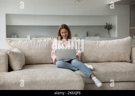 Hübsche Frau in legerer Kleidung sitzt auf dem Sofa mit Laptop Stockfoto