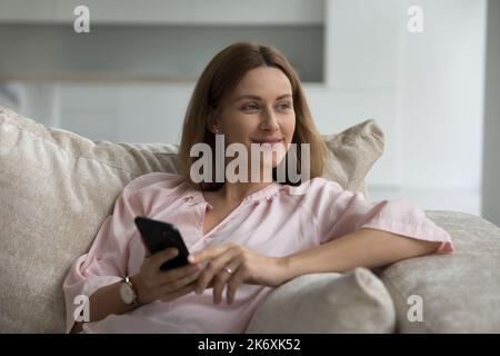 Frau schaut zur Seite sitzt auf dem Sofa mit Smartphone in den Händen Stockfoto