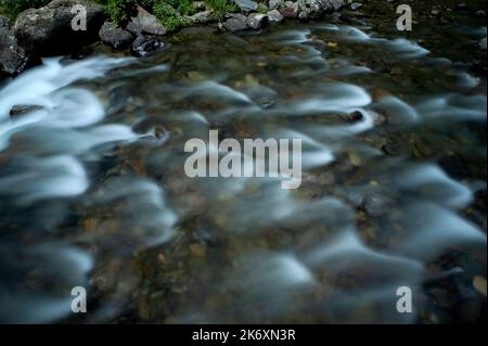 Die zeitbelichtende Fotografie verwischt die plätschernde Oberfläche von Andorras Valira del Nord, einem schnell fließenden Fluss, der hoch in den östlichen Pyrenäen aufsteigt und schließlich in die Gran Valira, den größten Wasserlauf des Fürstentums, mündet. Blick auf das Flussbett neben der Pont d’Ordino oder der Pont de l’Estarell, eine Buckelbrücke aus dem 15.. Jahrhundert, die 1980 nördlich von Llorts im Tal von Ordino umgebaut wurde. Stockfoto