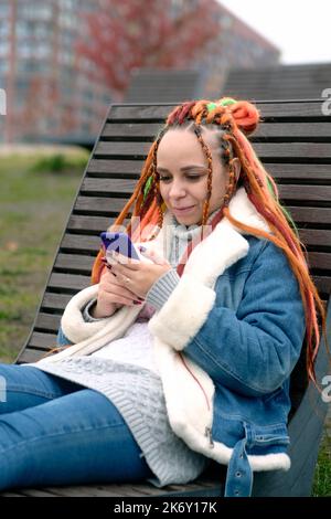 Junge Frau mit Dreadlocks in warmer Kleidung, die im Stadtpark auf einer hölzernen Sonnenliege auf dem Mobiltelefon surft. Sorglose Hündin mit farbenfroher Frisur usi Stockfoto