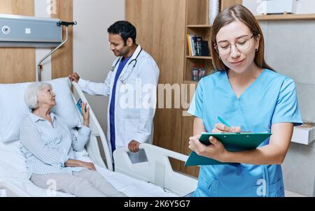 Lächelnde ältere Patientin, die mit ihrem Arzt im Krankenhauszimmer spricht, während die Krankenschwester Notizen in der Krankenakte macht. Medizinische Betreuung von Senioren Stockfoto