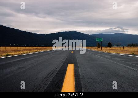 Leere Straße in Amerika an einem bewölkten Tag. Leere Straße in Kalifornien durch ein gelbes Feld mit Blick auf die Berge in der Ferne Stockfoto