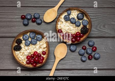 Ganze Hafer, Blaubeeren und Preiselbeeren in Kokosnussschalen. Zwei Holzlöffel und Beeren auf dem Tisch. Flach liegend. Dunkler Holzhintergrund. Stockfoto