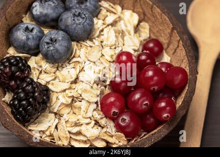 Ganze Hafer, Blaubeeren und Preiselbeeren in Kokosnussschalen. Holzlöffel auf dem Tisch. Nahaufnahme. Dunkler Holzhintergrund. Stockfoto