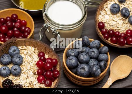 Frühstückscerealien. Ungekochte Haferflocken mit Beeren in Kokosnussschalen. Heidelbeeren und Preiselbeeren in Holzschüssel. Joghurt im Glas. Draufsicht. Stockfoto