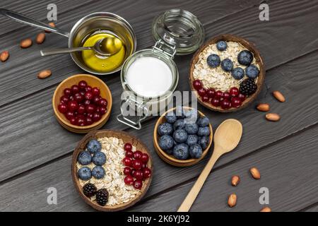 Ungekochte Haferflocken mit Beeren. Joghurt im Glas, Honig in Metallschüssel. Preiselbeeren und Heidelbeeren in Holzschüsseln. Muttern auf dem Tisch. Flach liegend. Dunkles Holz b Stockfoto