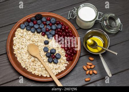 Ungekochte Haferflocken mit Beeren und Holzlöffel auf Keramikplatte. Joghurt im Glas, Honig in Metallschüssel. Flach liegend. Dunkler Holzhintergrund. Stockfoto