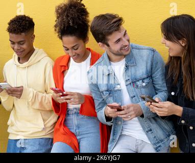 Eine Gruppe von verschiedenen, tausendjährigen jungen Freunden, die lächeln, während sie auf das Mobiltelefon über der gelben Wand blicken Stockfoto