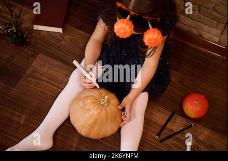 Kleines Mädchen in einem schwarzen Kleid und Kürbisse auf einem Reifen, das zu Hause eine Jack-O-Laterne macht und ein gruseliges Gesicht auf einen Kürbis malt Stockfoto