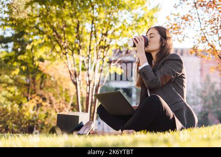 Ganzkörperfoto Frau, die mit einem Laptop im Internet surft, sich auf grünem Rasen im Park ausruht, Kaffee trinkt, sich im Freien entspannt. Lifestyle, Kaffee Stockfoto