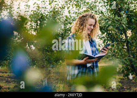 Frau Agronomin hält ein Telefon und Notizblock stehen in Obstgarten, Qualität Kontrolle der Produktion des Jahres. Landarbeiter. Landwirtschaft Stockfoto
