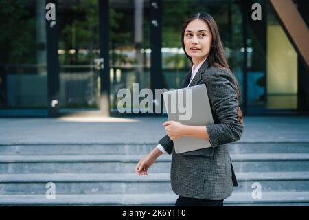 Erfolgreiche Geschäftsfrau in Jacke, die während der Pause draußen mit einem Laptop steht. Geschäftsleute. Moderne Dame draußen Stockfoto