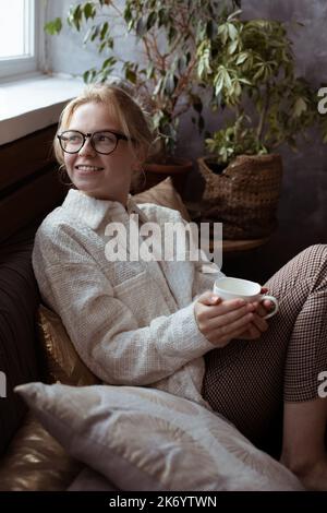 Vertikal positiv lächelnde blonde Frau in einer Brille auf gemütlichem Sofa in der Nähe von Zimmerpflanzen sitzen und Tasse halten, Kaffee oder Tee trinken Stockfoto