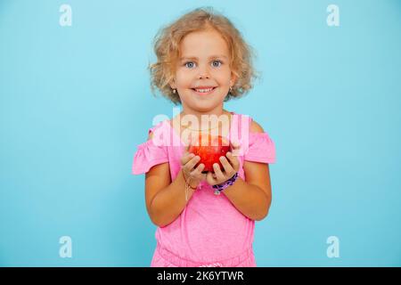 Kleines, kleines lächelndes, ausdrucksstarkes, positiv lockiges blondes Mädchen, das roten Saftapfel in den Händen auf blauem Blick hält. Gesundes Leben Stockfoto